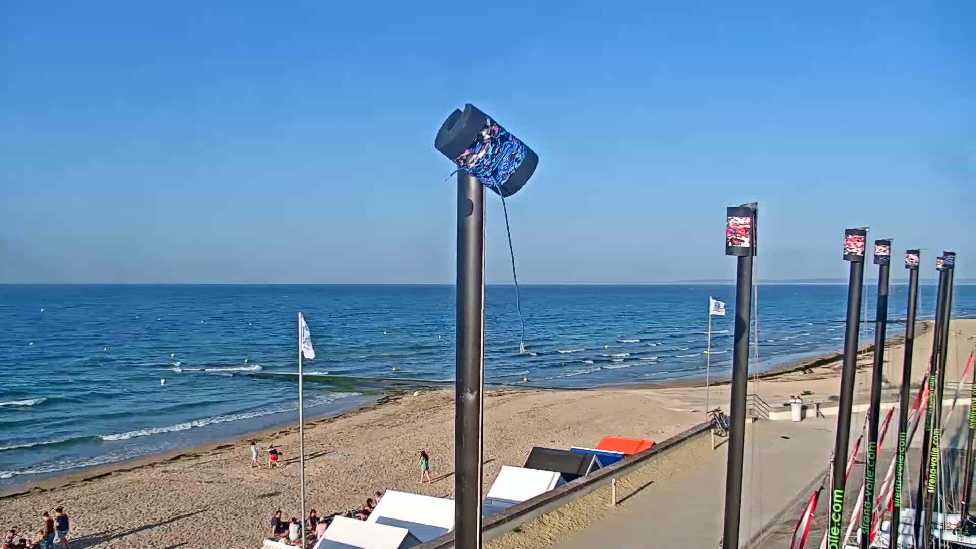 A sandy beach with calm ocean waves under a clear blue sky, featuring several beach cabanas and a few people enjoying the sunny day.