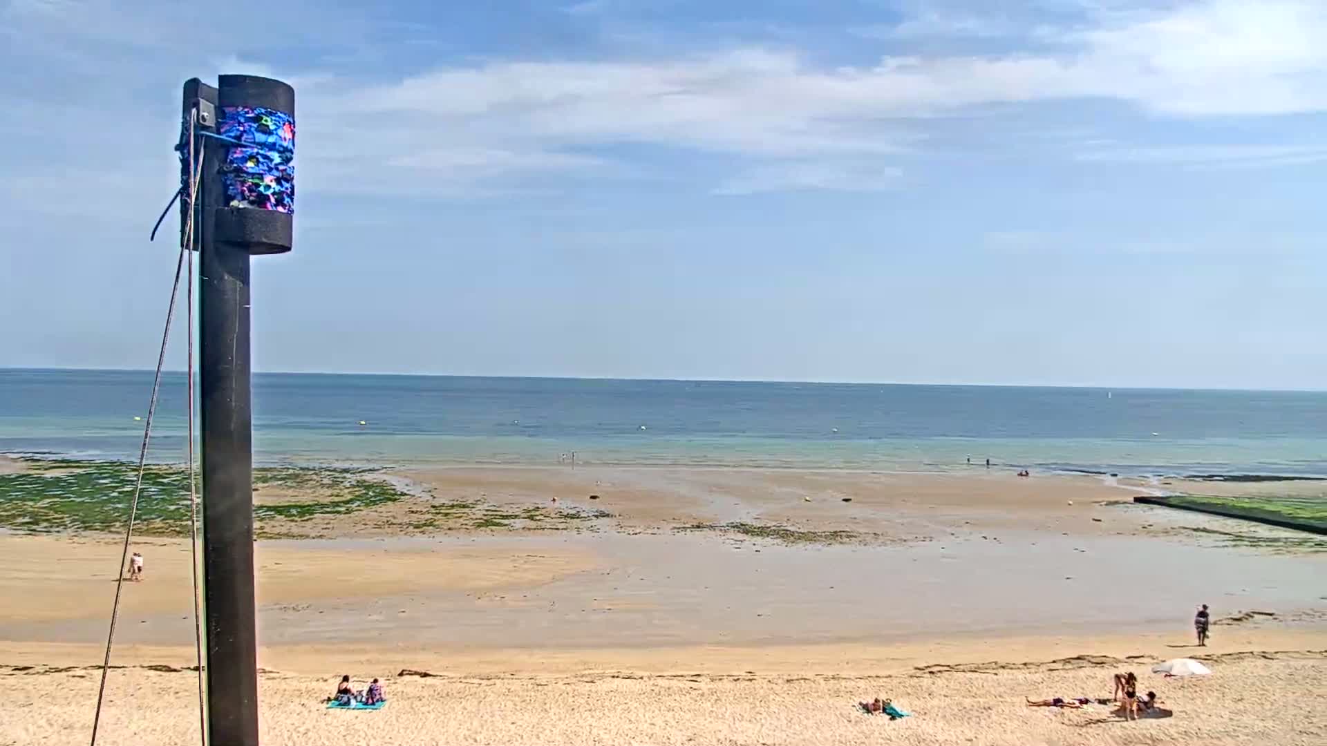 A sandy beach with a few people sunbathing and walking, a calm ocean under a mostly sunny sky, and a tall, slender pole with a decorated top in the foreground.