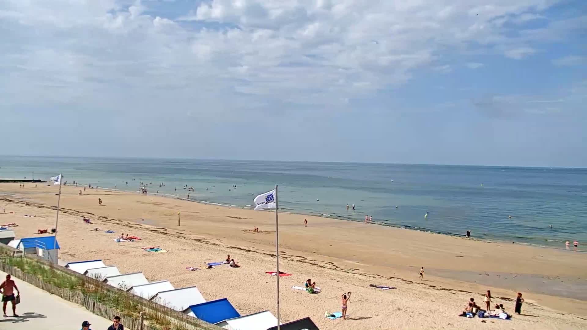 A sandy beach with scattered people sunbathing and swimming in a calm ocean under a partly cloudy sky.
