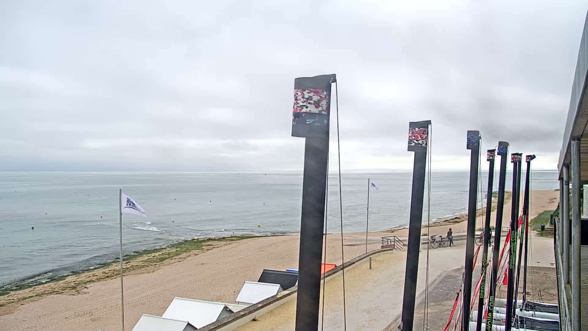 A sandy beach and calm ocean are visible under an overcast sky, with several tall masts and a few people near a building in the foreground.