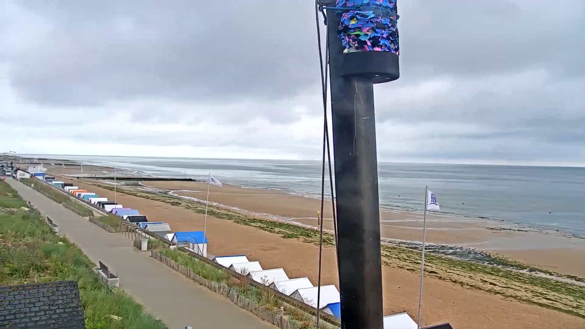 A beach with colorful beach huts and a paved walkway is seen under an overcast sky, overlooking a calm sea.