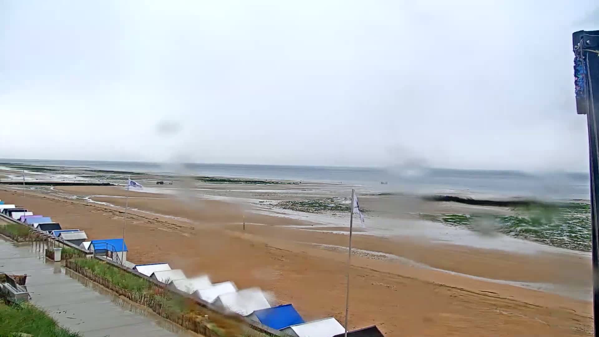 A wet sandy beach with rows of beach huts is visible under an overcast sky, with a calm sea in the background.