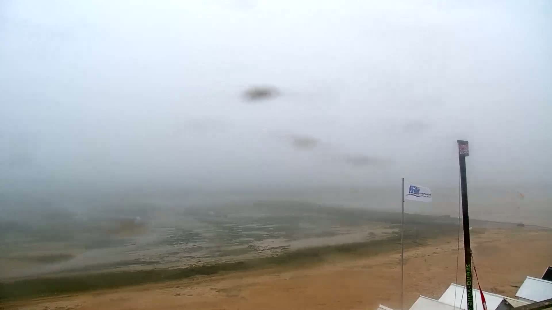 A foggy beach scene shows a sandy shoreline and a body of water in the distance, with a tall mast and some small structures visible in the foreground.