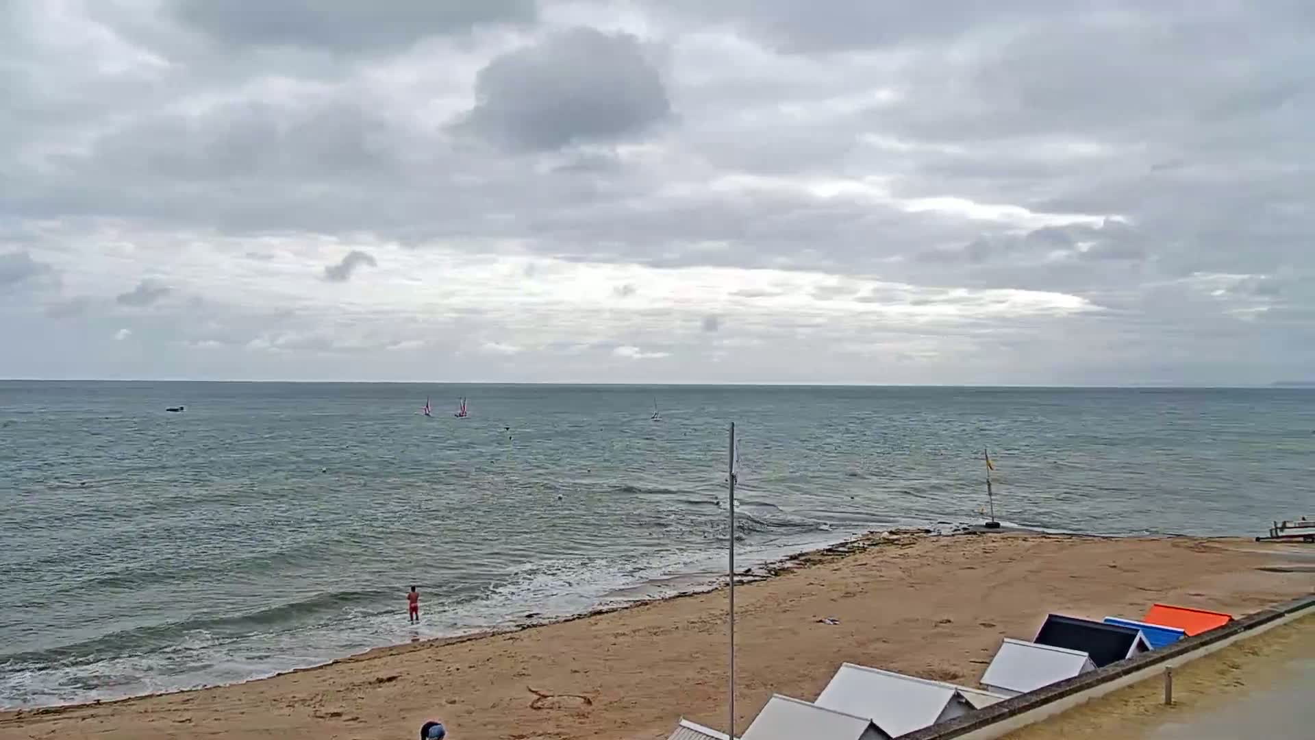 A sandy beach with several small beach huts slopes down to a calm sea under a cloudy sky, with a few sailboats visible on the water.