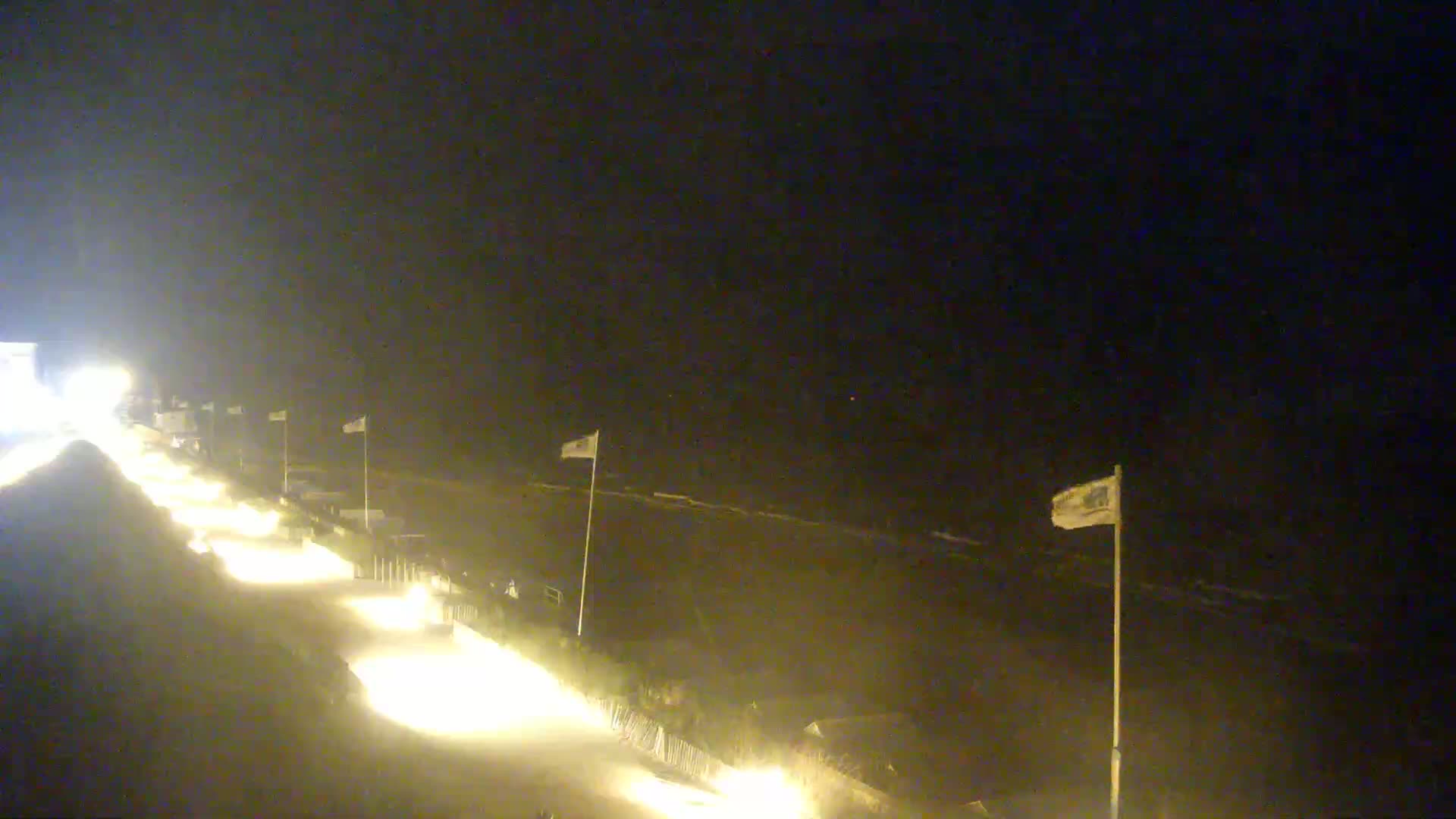 A nighttime beach scene shows a dimly lit boardwalk with several flags and a dark ocean with small waves under a clear sky.