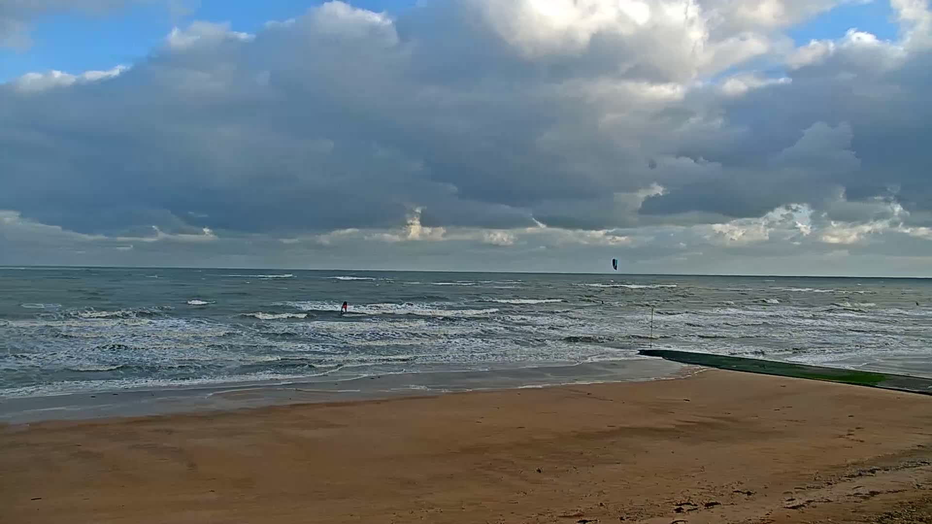 On an overcast day with patches of blue sky, a windsurfer and a kitesurfer navigate the choppy sea off a sandy beach, with a dark pier extending into the water on the right.