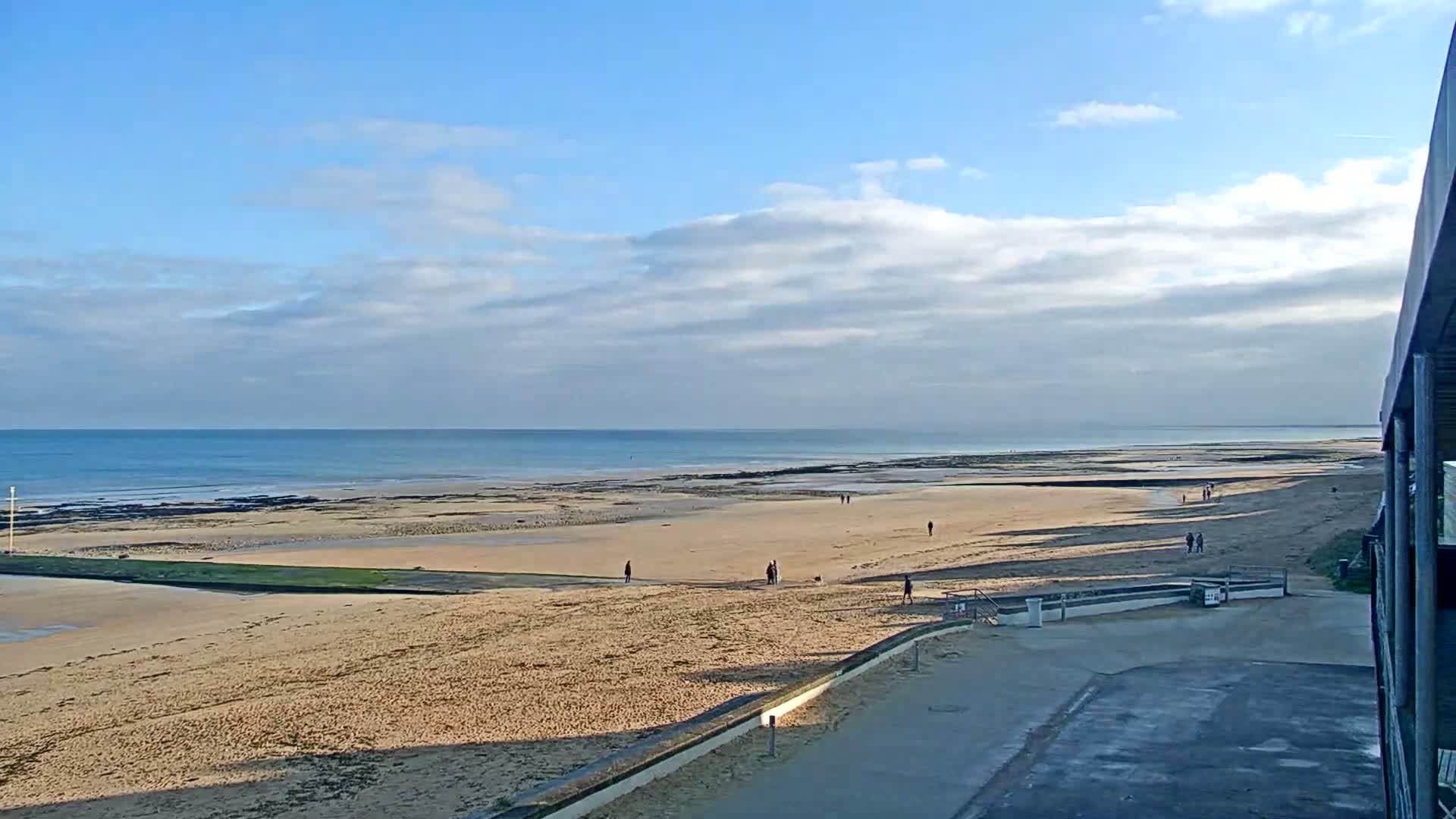 On an overcast day with patches of blue sky, a windsurfer and a kitesurfer navigate the choppy sea off a sandy beach, with a dark pier extending into the water on the right.
