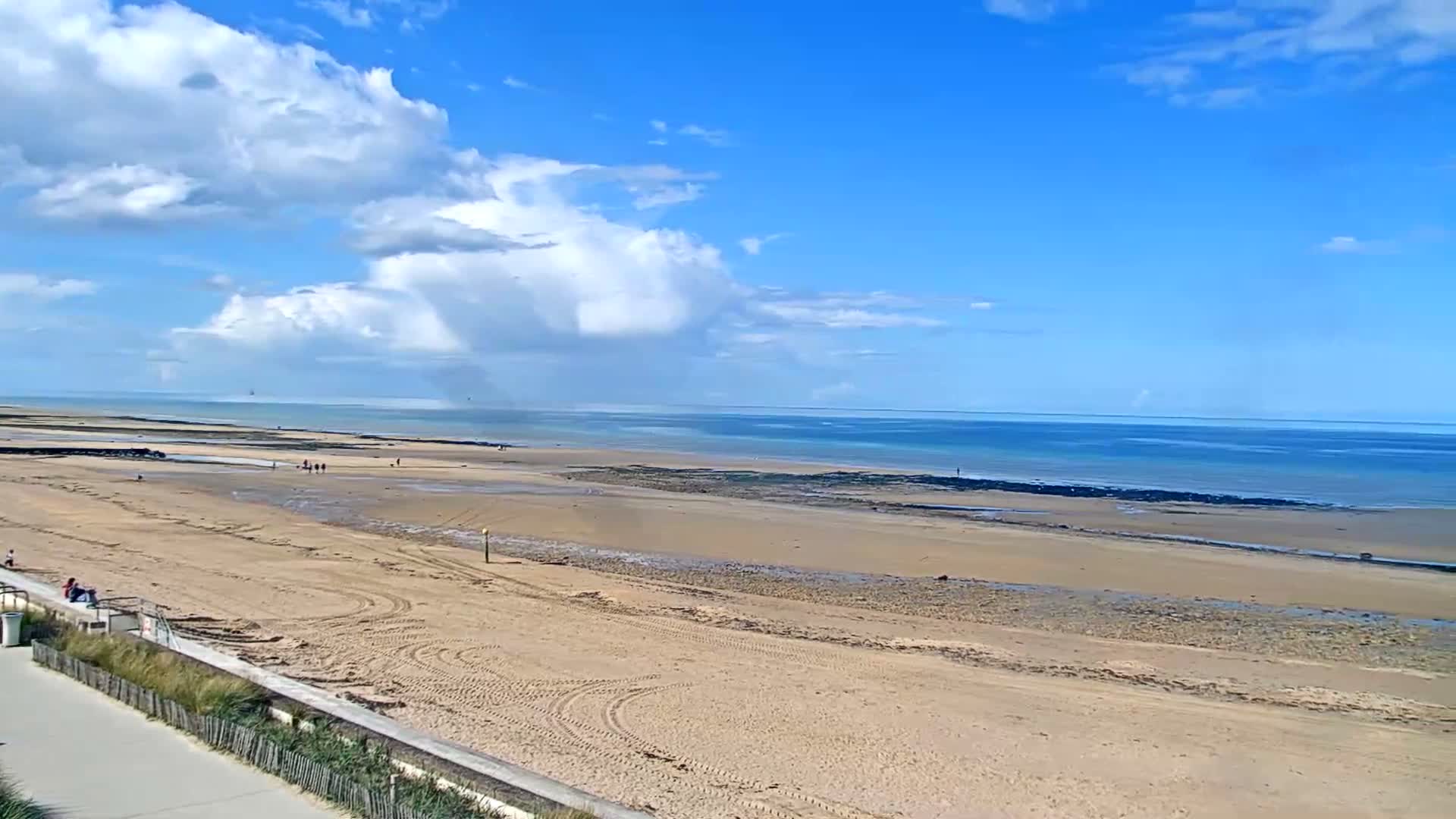 On an overcast day with patches of blue sky, a windsurfer and a kitesurfer navigate the choppy sea off a sandy beach, with a dark pier extending into the water on the right.