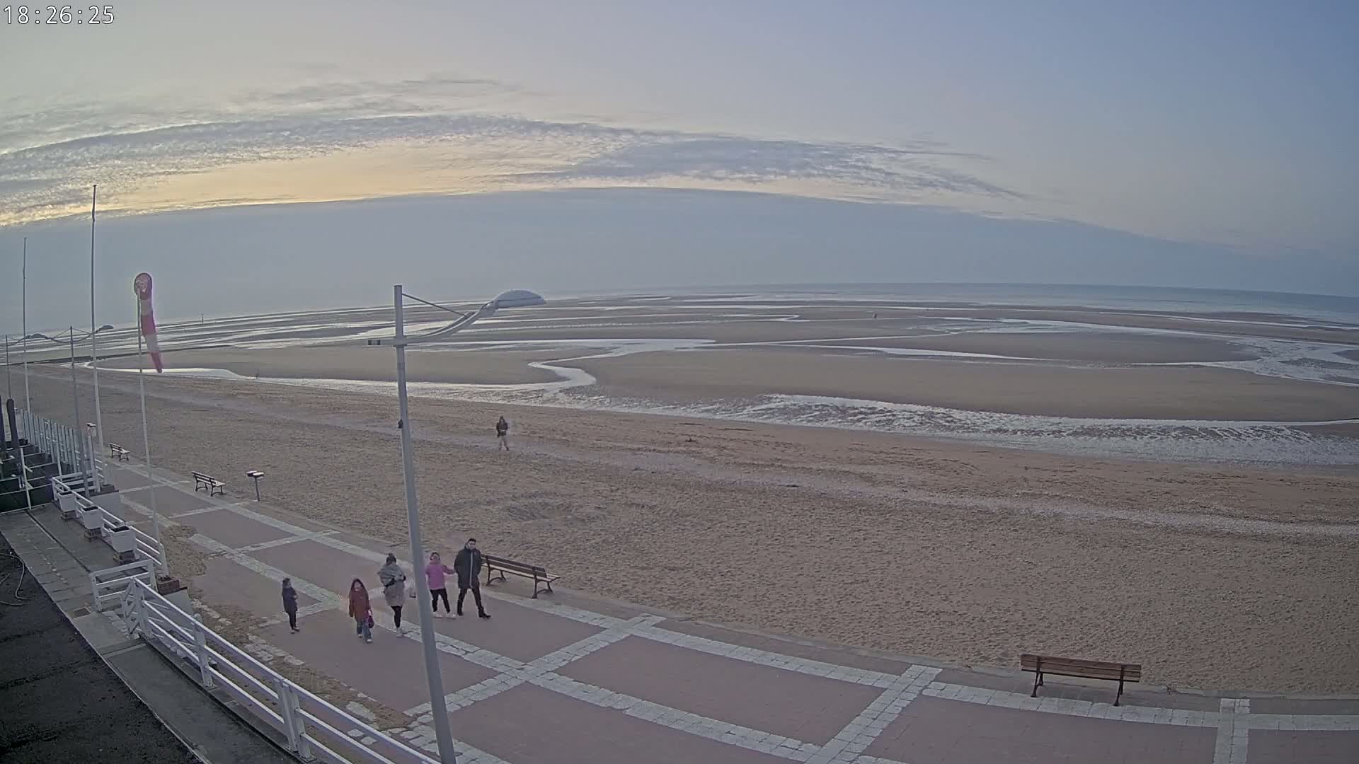 A wide sandy beach stretches to a calm ocean under a pale, cloudy sky, with several people walking along a paved walkway near the shoreline.
