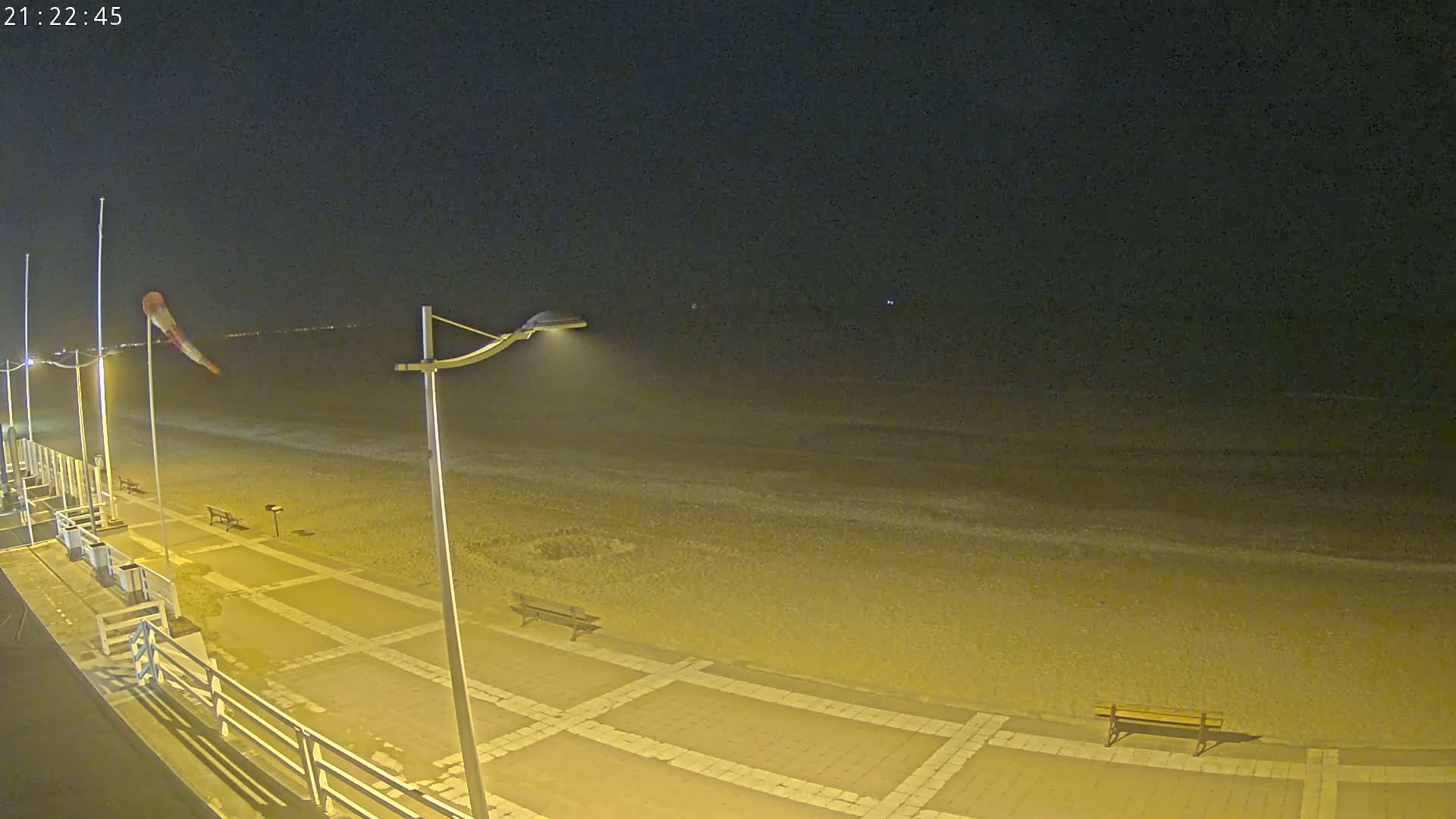 A nighttime view of a beach with a paved walkway, benches, and lamps, under a dark sky.
