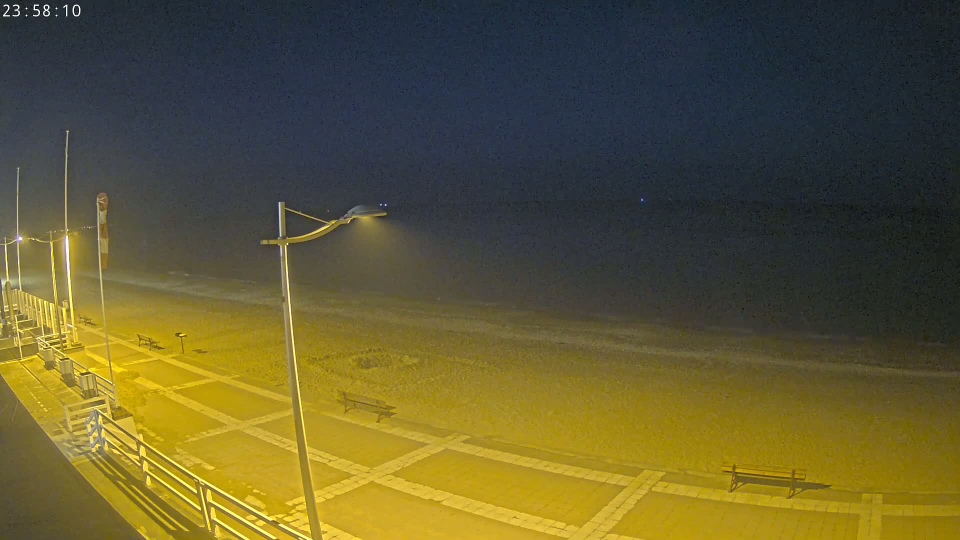 A nighttime view of a beach with a paved walkway, benches, and lights, under a dark sky.