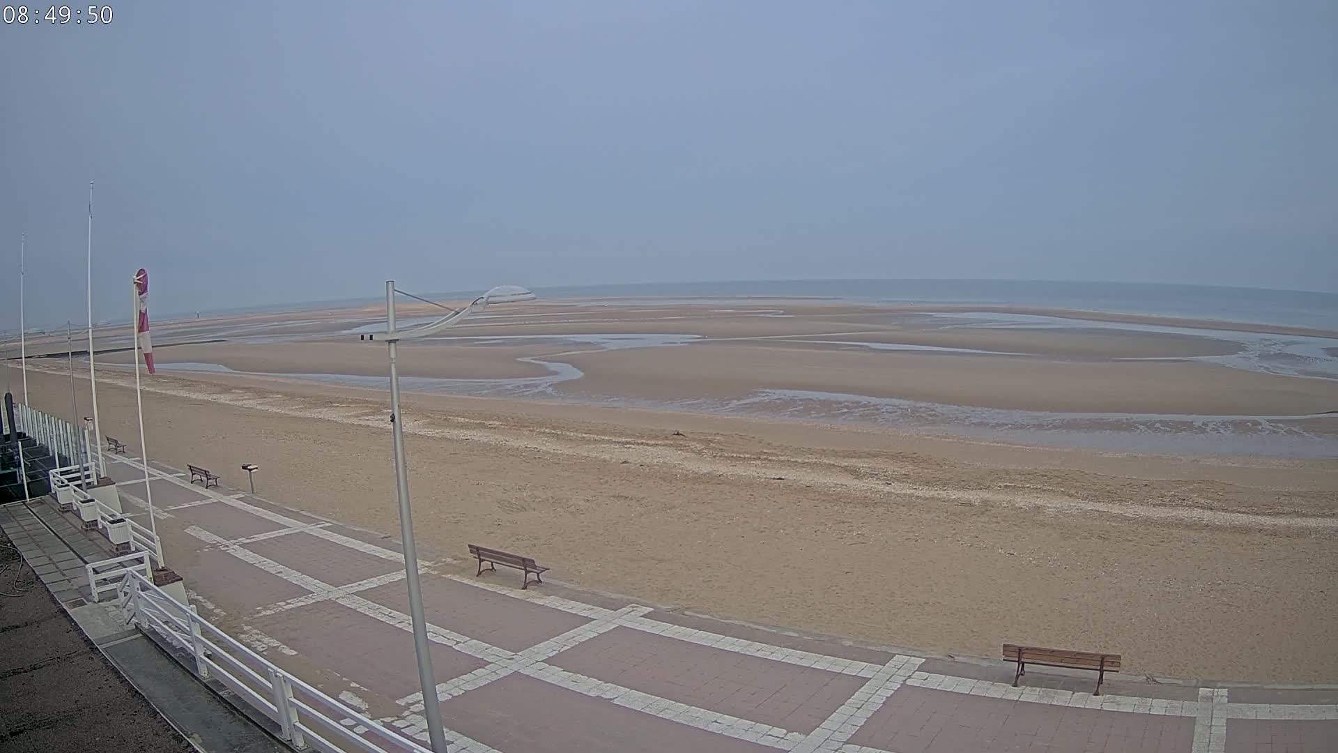 A wide sandy beach with low tide, a paved walkway with benches, and a windsock on a mostly cloudy day.