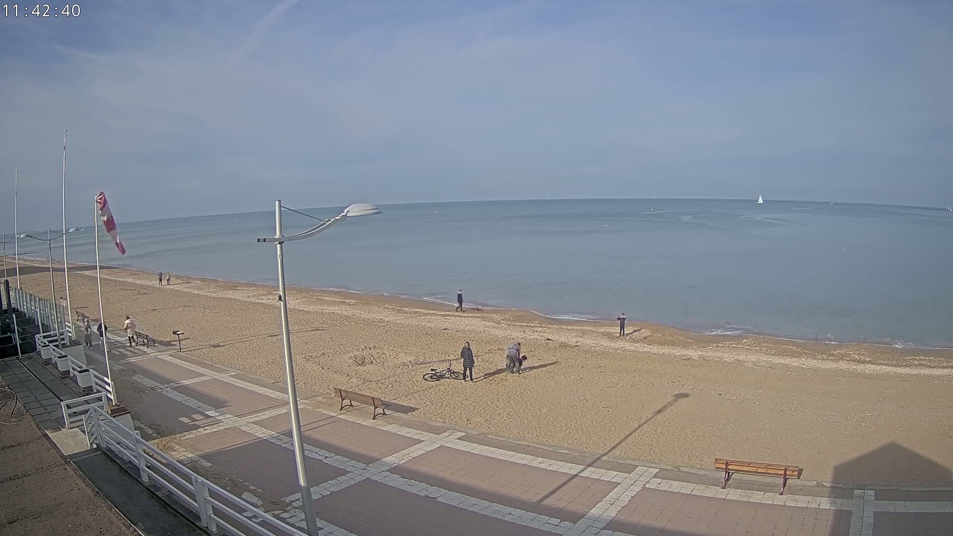 A sandy beach with a paved walkway and several people is visible, overlooking a calm sea on a sunny day.