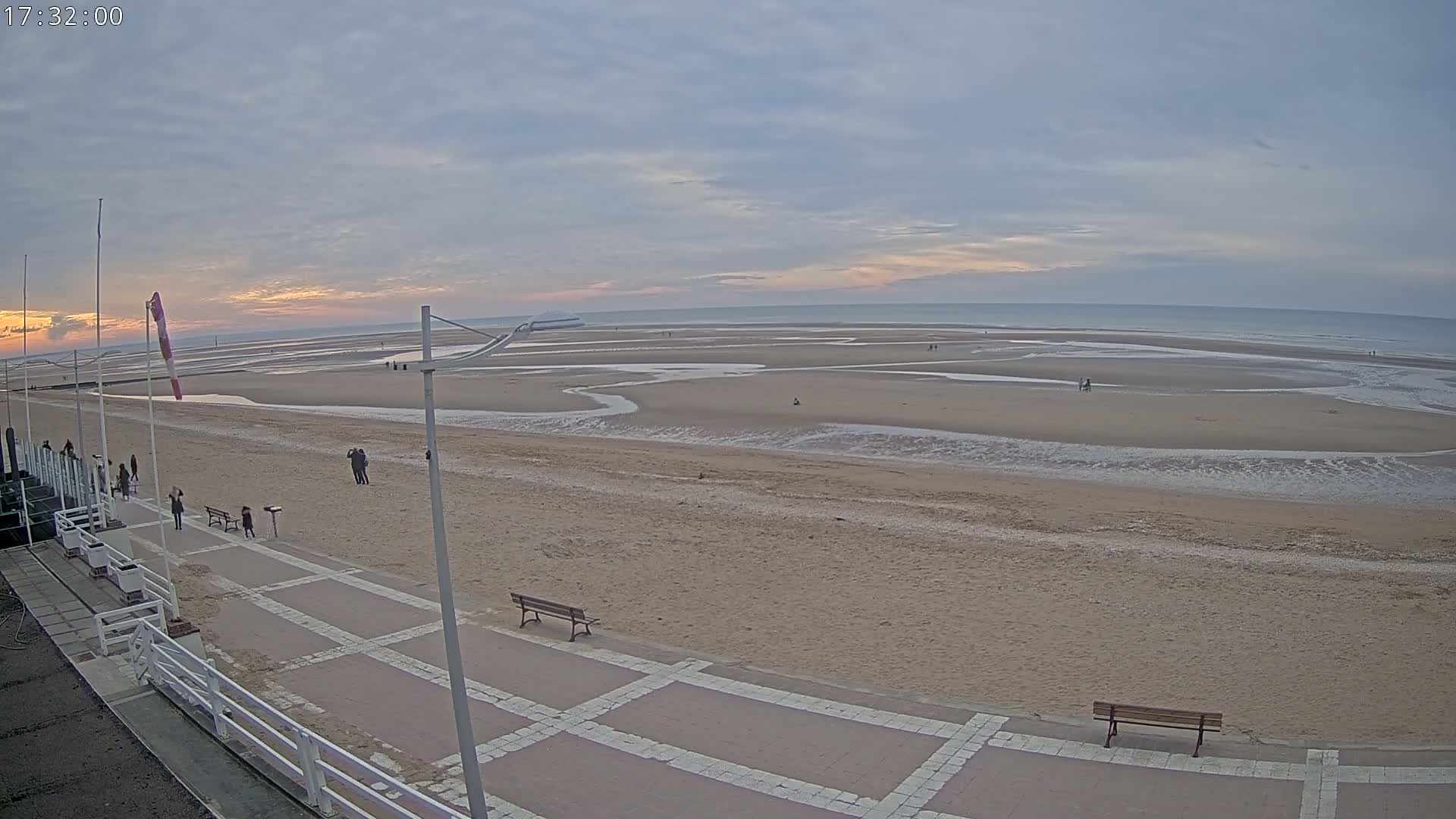 A wide shot of a mostly empty sandy beach at sunset, with a paved walkway and benches visible in the foreground, under a cloudy sky.