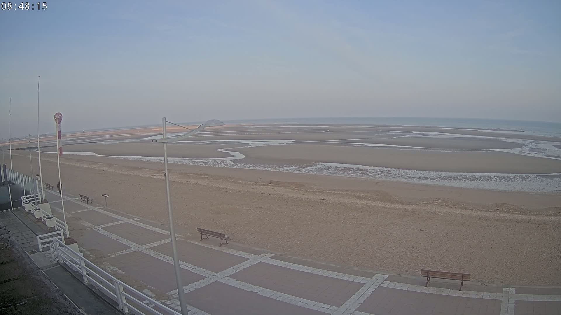 A mostly empty sandy beach stretches to a calm ocean under a hazy sky, with a paved walkway and benches visible in the foreground.