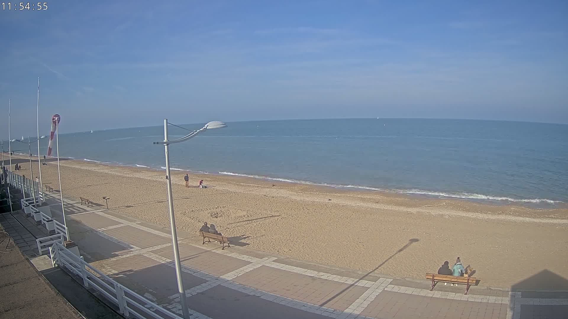 A sandy beach with a paved walkway, several people, and calm ocean under a clear blue sky.