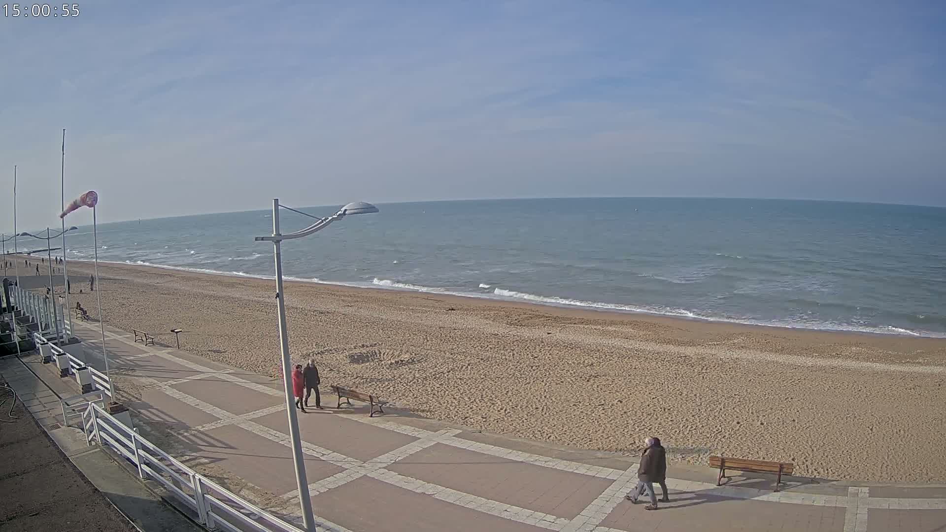 A mostly sunny day shows a wide sandy beach with a paved walkway, a few people strolling, and calm waves lapping the shore.