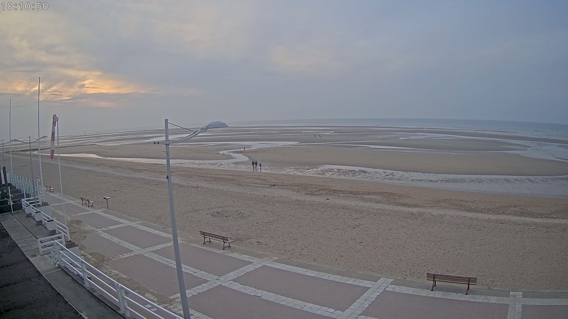 A wide sandy beach with a paved walkway, benches, and a few people walking stretches to a calm ocean under a cloudy, dusky sky.