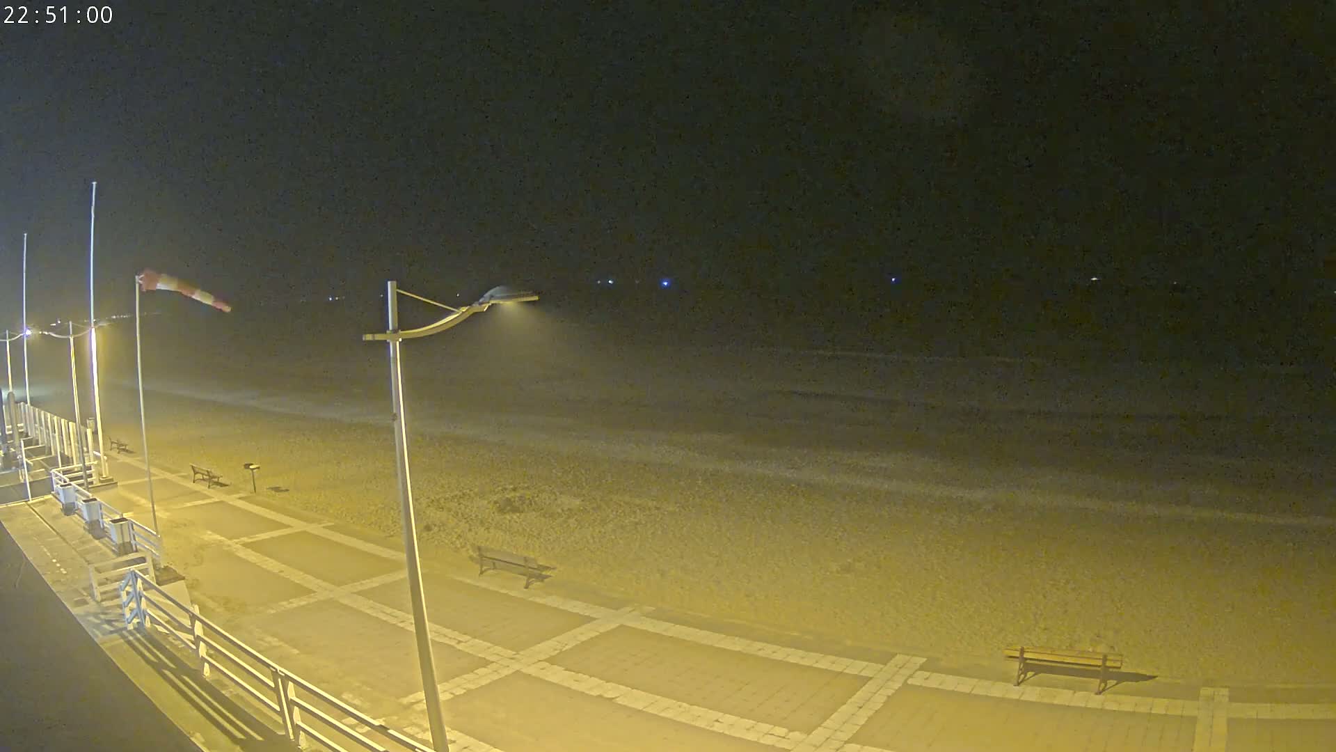 A nighttime view of a beach with a paved walkway, benches, and streetlights, under windy conditions.