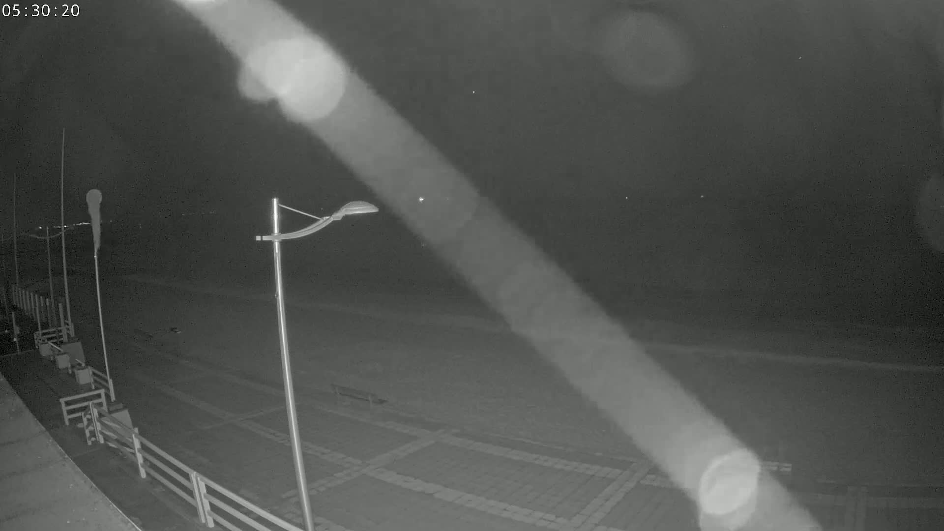 A nighttime, low-light view of a paved beachfront area with a lamppost and railings, under a dark sky.