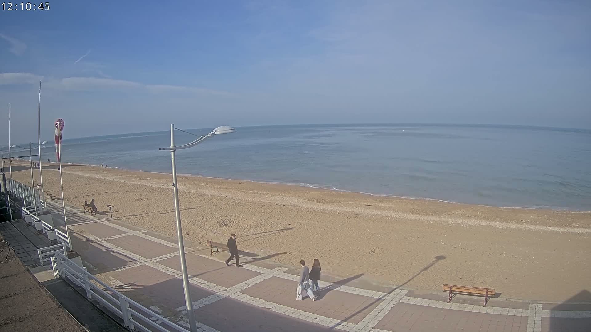 A sandy beach stretches alongside a paved walkway where several people stroll under a clear, bright sky.