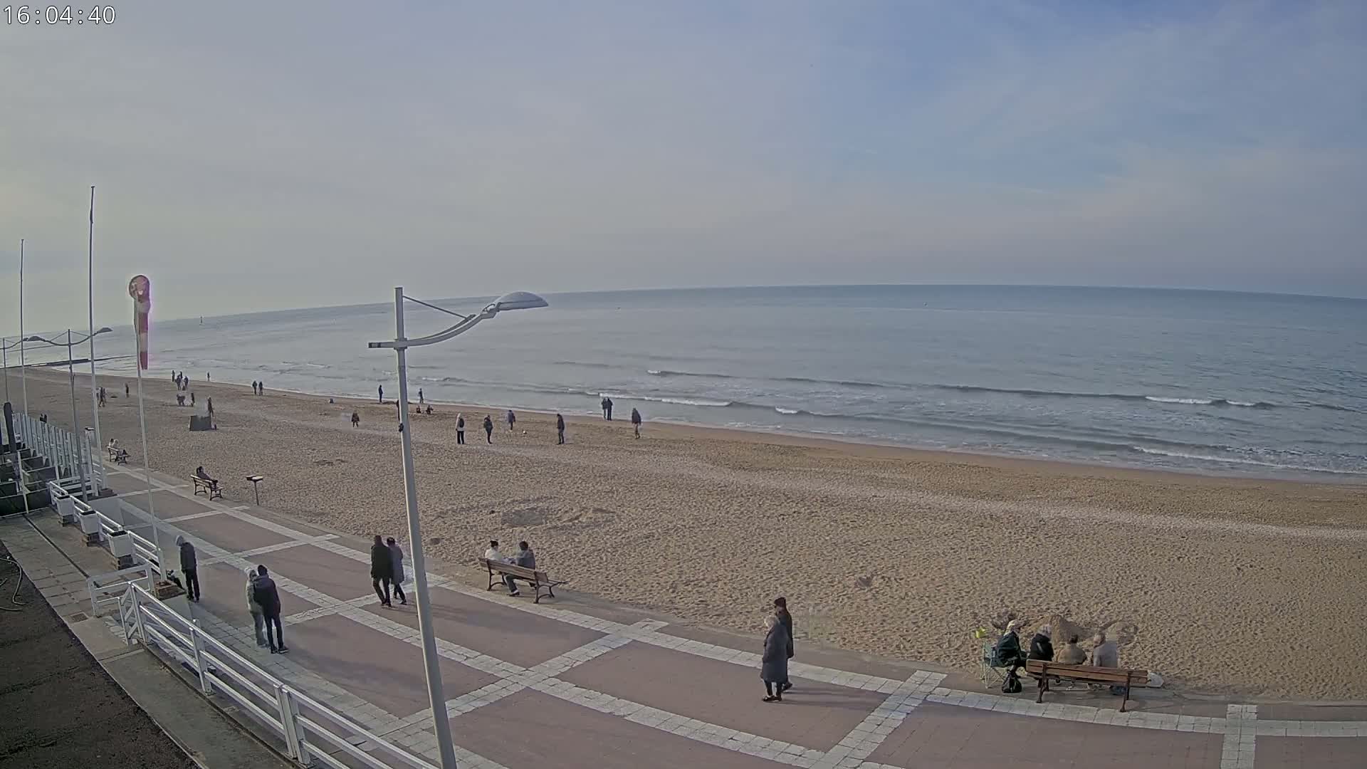 A mostly sunny day at a sandy beach with a promenade, where several people are walking and sitting.