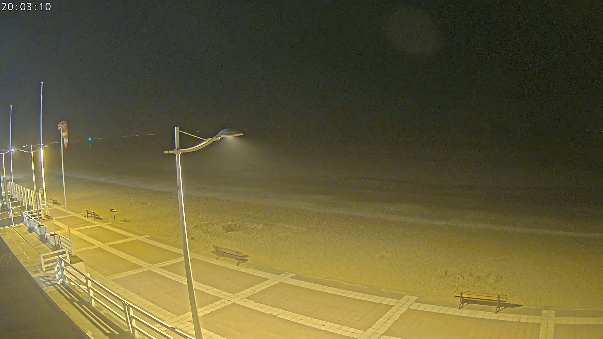 A nighttime view of a beach with a paved walkway, benches, and lampposts, under a dark sky.