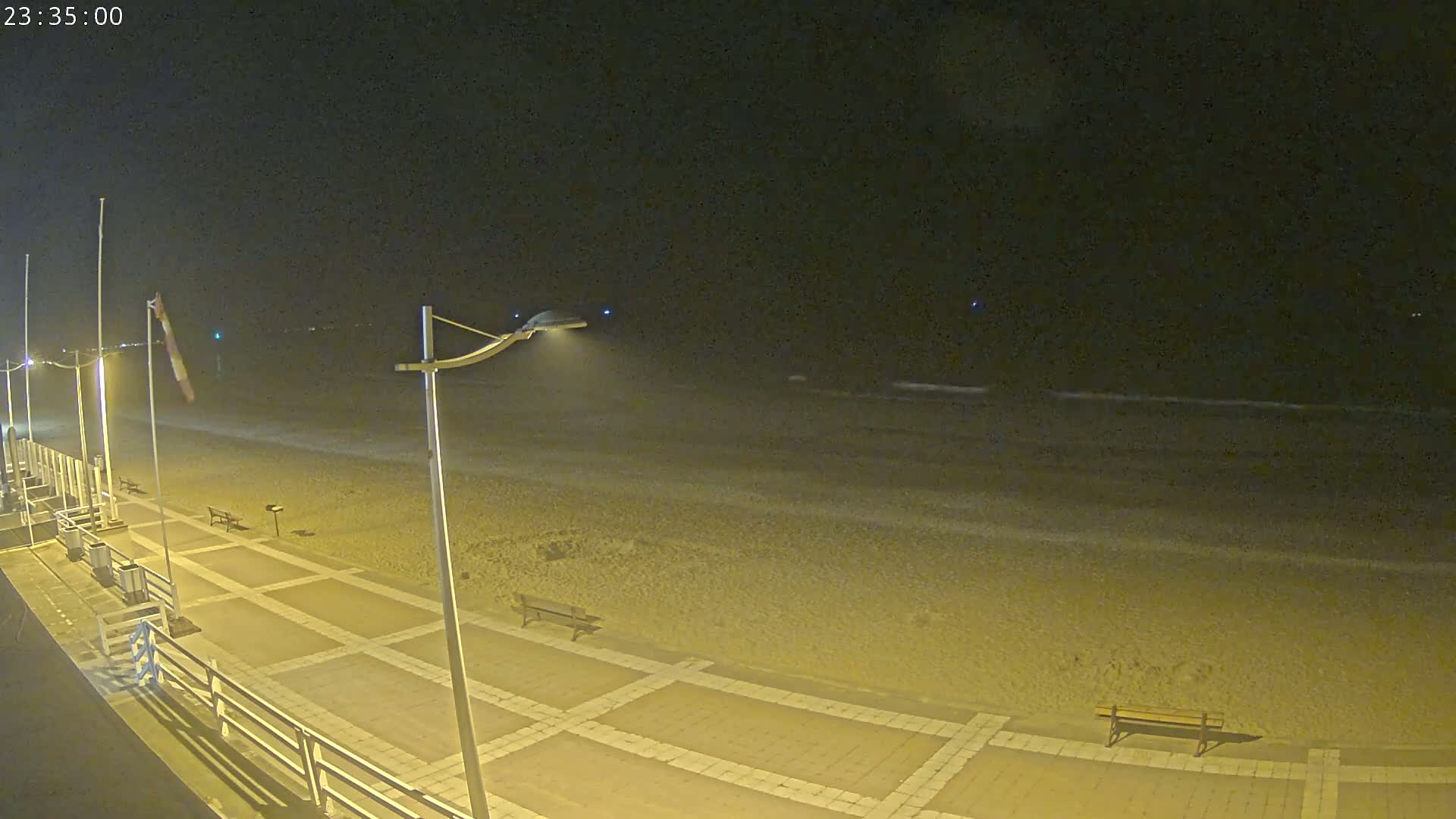 A nighttime view of a mostly empty beach with a paved walkway, benches, and streetlights.