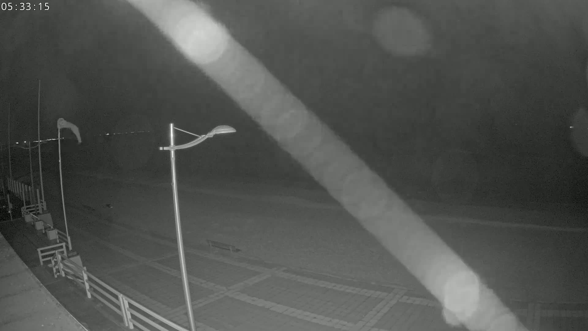 A nighttime, low-light image shows a paved area near a body of water, with a streetlight, flags waving in the wind, and a fence.