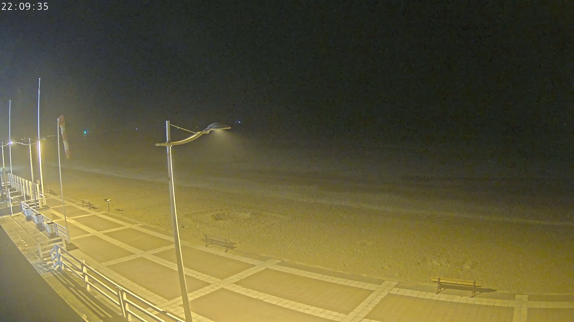 A nighttime view of a paved beachfront walkway with benches, lit by streetlights, overlooking a dark, gently lapping ocean under a clear sky.