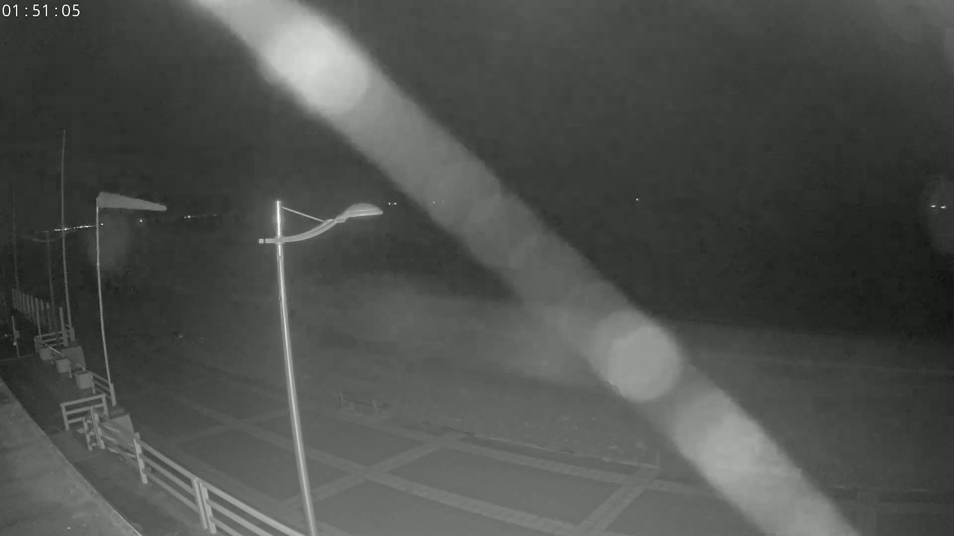 A nighttime, low-light view shows an empty paved area with a lamppost and windsock, possibly near a body of water.