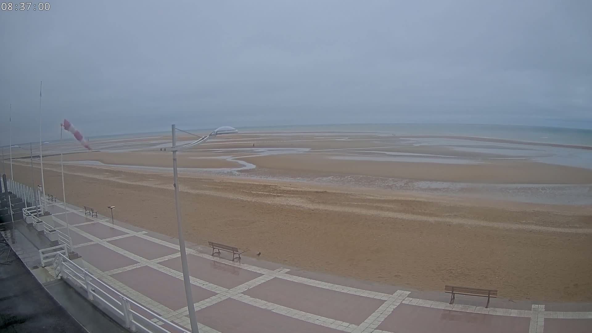 A wide, sandy beach stretches to a calm, grey sea under an overcast sky, with a paved walkway and benches visible in the foreground.