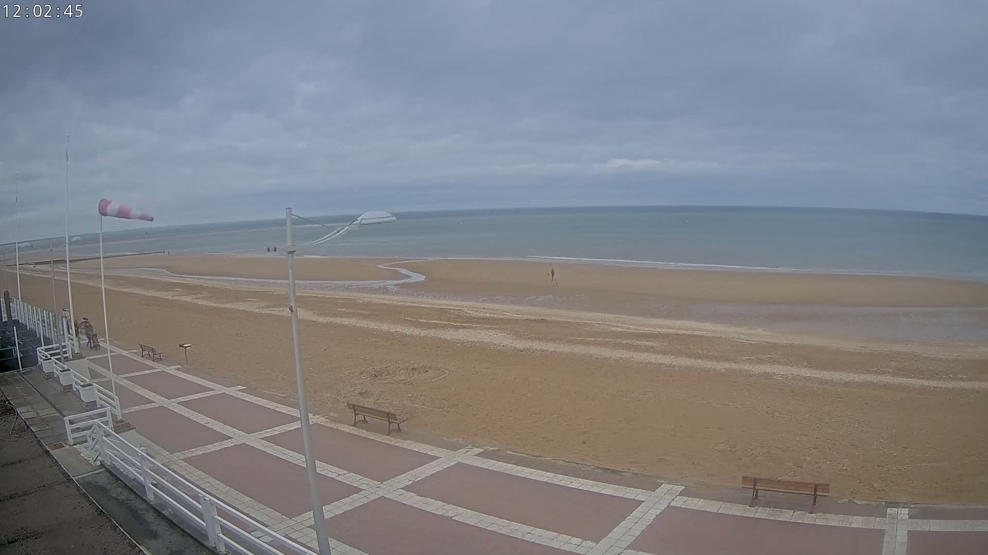 A mostly-empty sandy beach stretches to a calm, gray sea under an overcast sky, with a paved boardwalk and benches visible in the foreground.