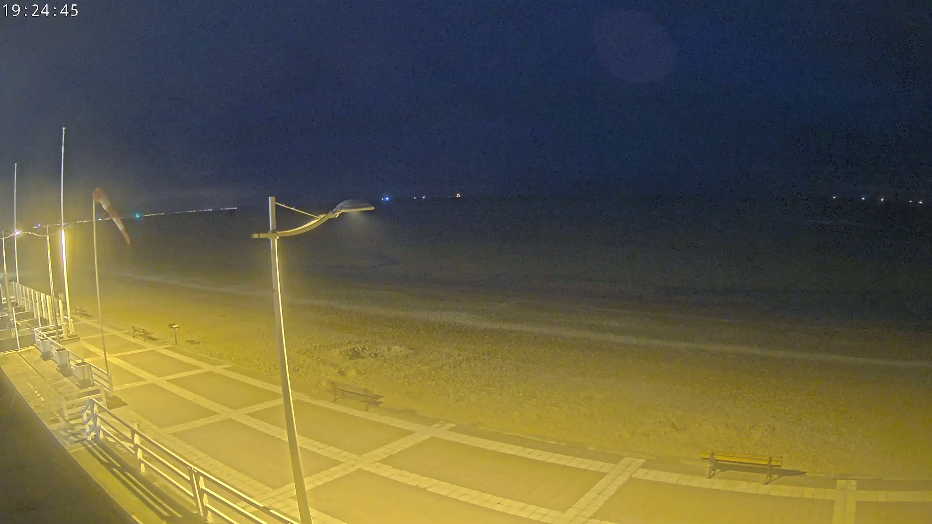 A nighttime view of a beach promenade with a dark sky, illuminated by streetlights, showing a mostly empty sandy beach and calm water.