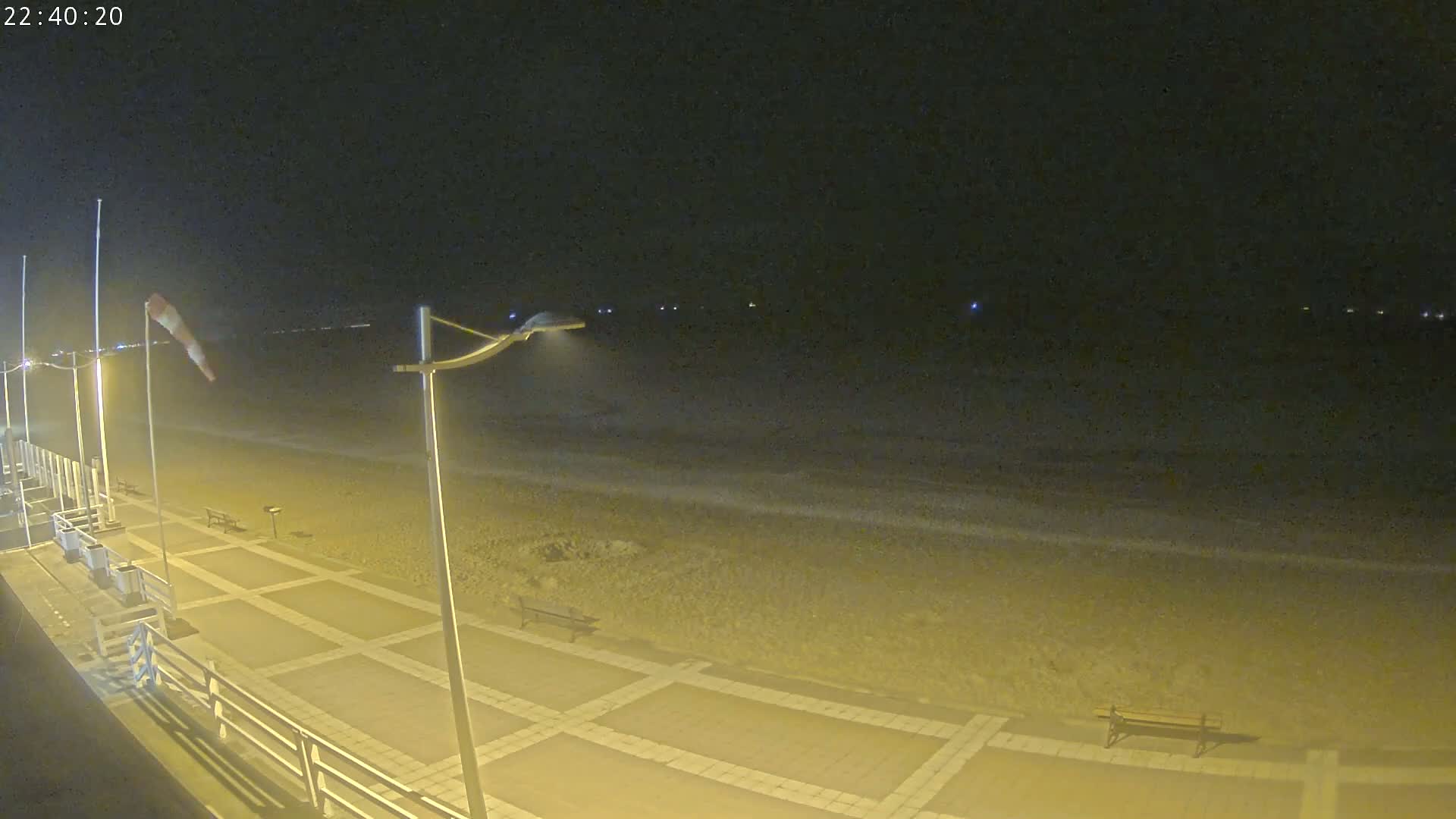 A paved beachfront walkway at night with a windsock indicating wind, gentle waves lapping the shore under a dark sky.