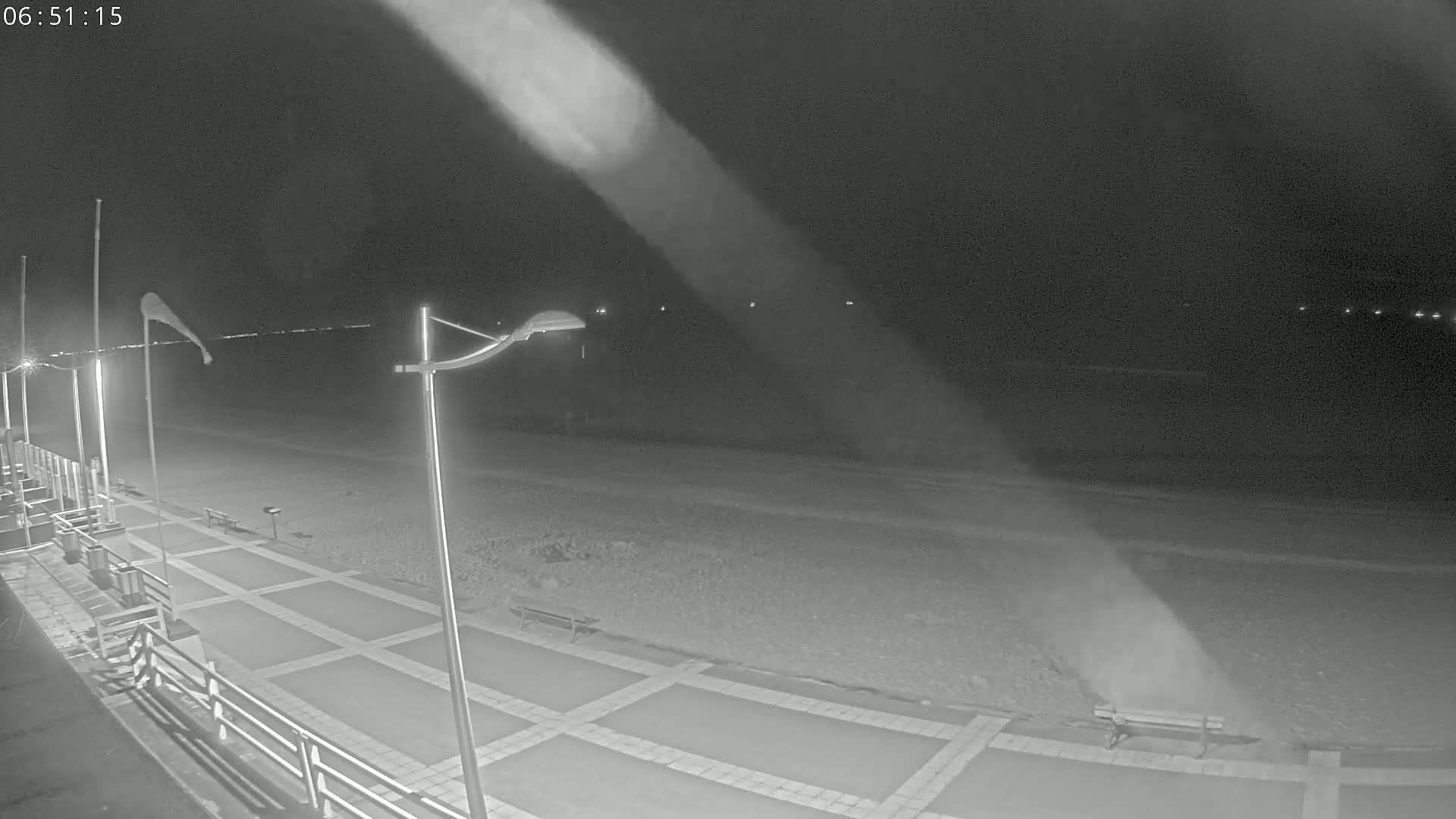 A nighttime view of a paved walkway next to a beach, with wind blowing sand across the beach and flags flapping on poles.
