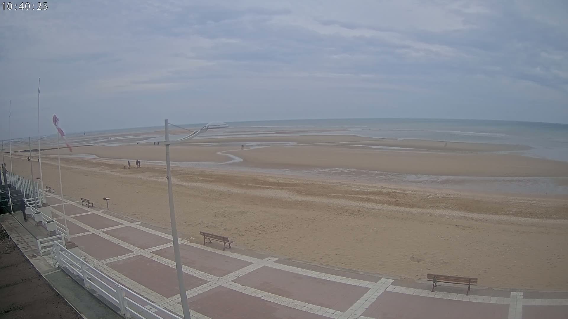 A wide sandy beach stretches to a calm sea under an overcast sky, with a paved walkway and benches visible in the foreground.