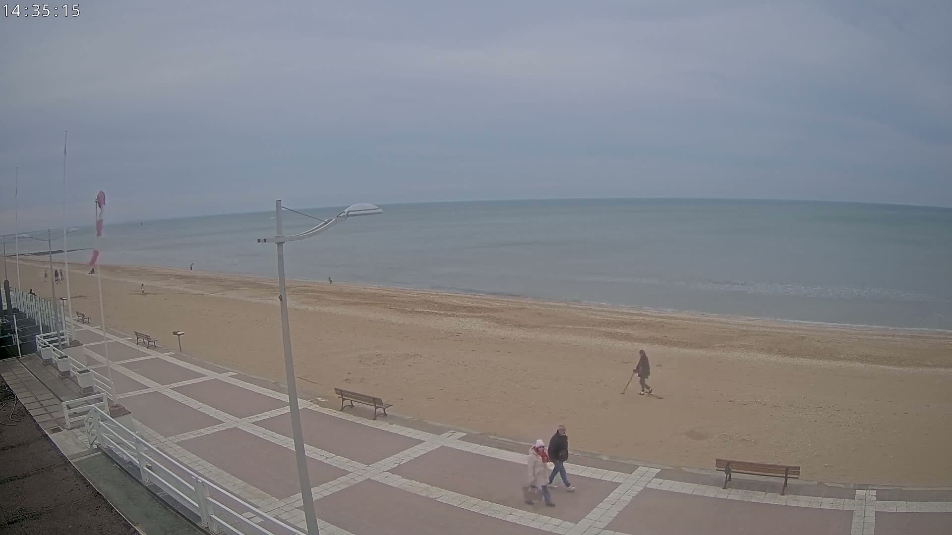 A mostly overcast day shows a sandy beach with a few people walking along the shore and promenade.