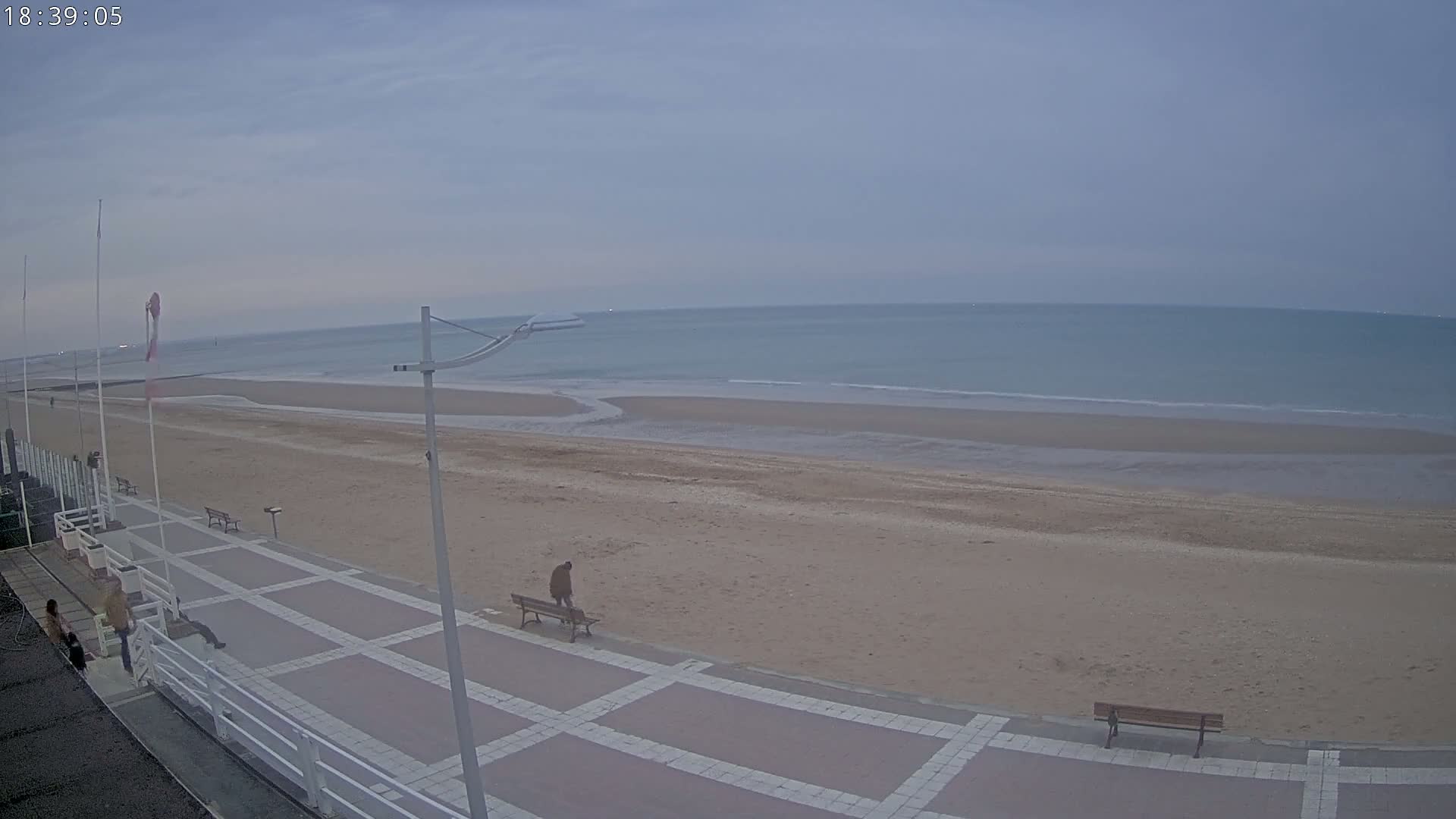 A mostly empty sandy beach and paved walkway are visible under an overcast sky, with a calm sea in the background.
