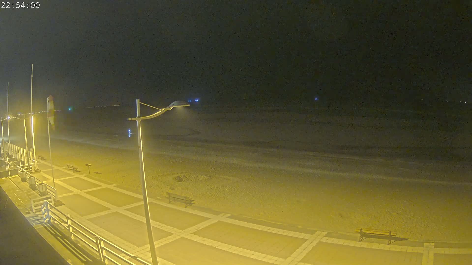 A nighttime view of a paved beach walkway next to a dark sandy beach under a clear, dark sky.