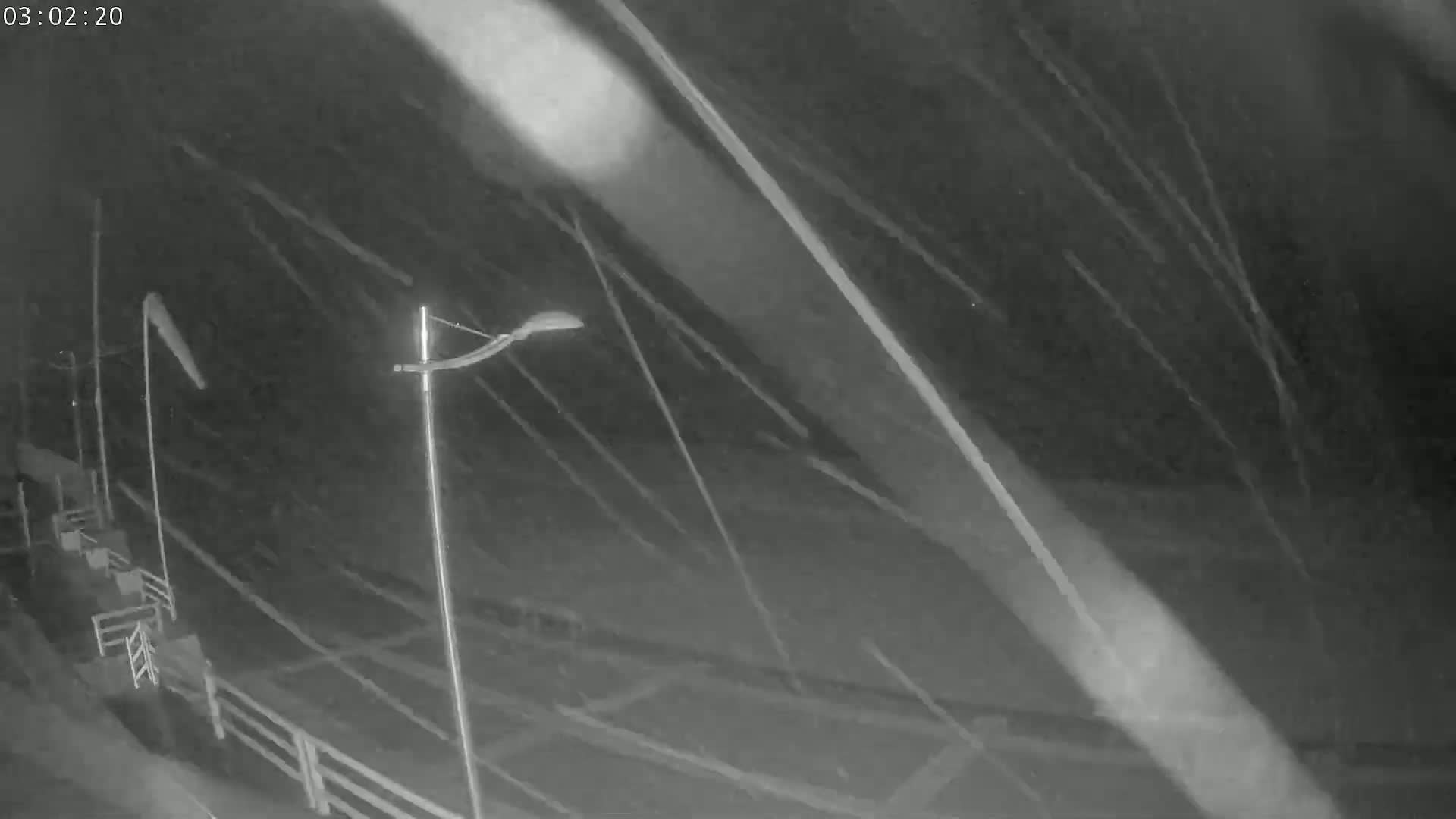 A nighttime, snowy scene shows a roadway with poles and some fencing, viewed from an elevated perspective.