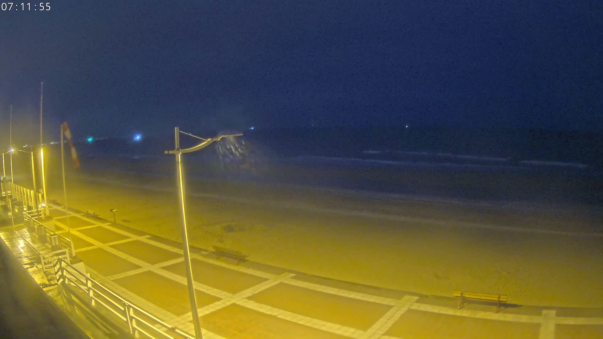 A nighttime view of a beach at night with mildly wavy water under a dark sky, showing a paved walkway with lights and a bench.