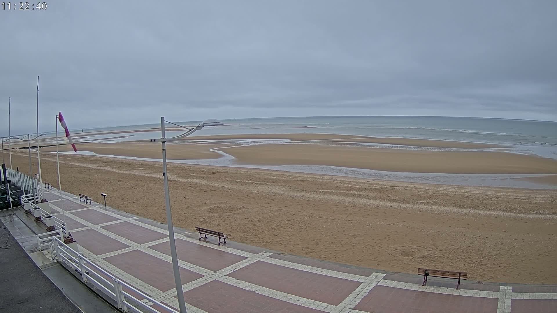 A paved walkway with benches overlooks a wide, sandy beach and calm ocean under an overcast sky.