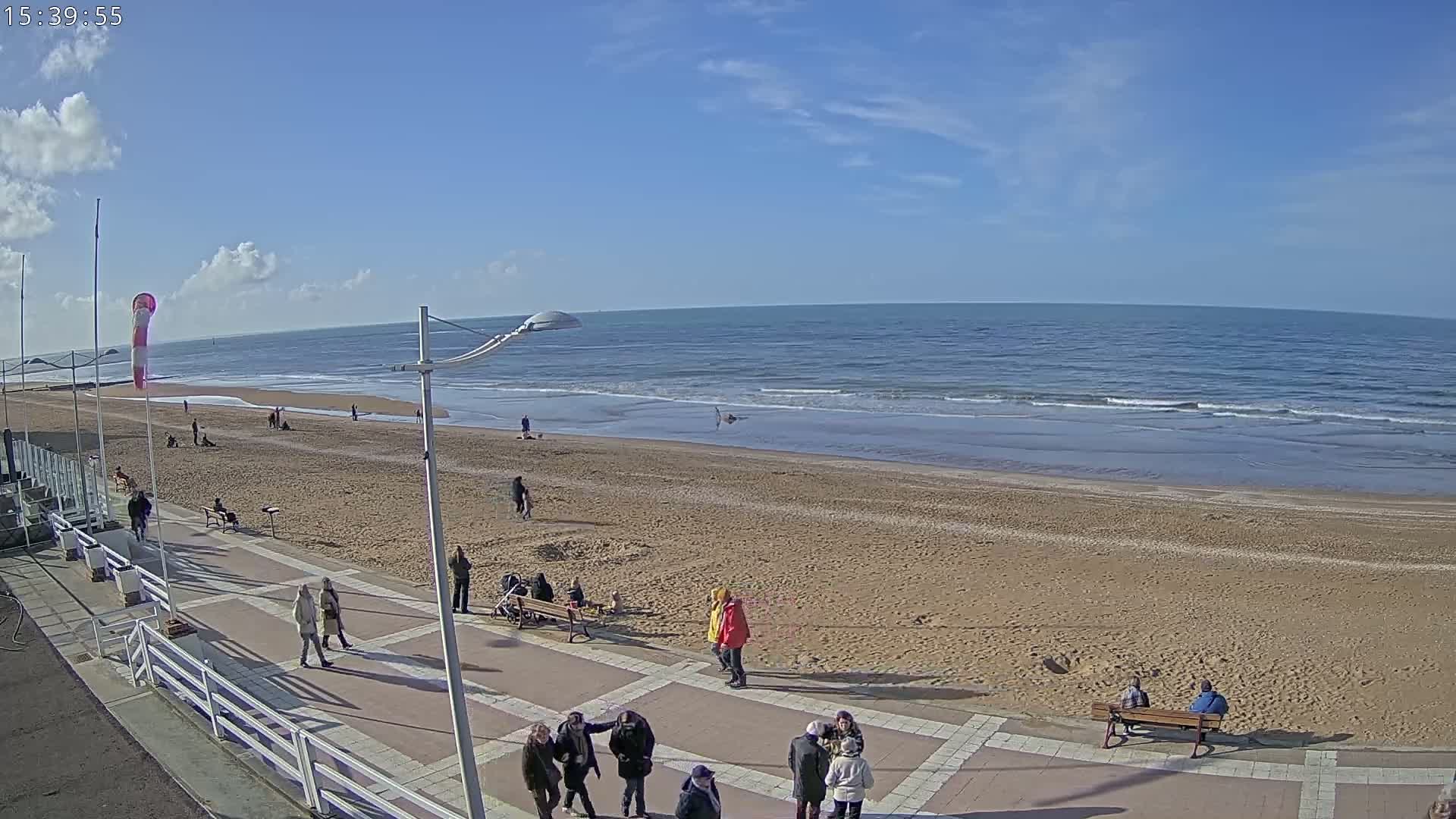 A sandy beach with a few people walking along a paved walkway, under a mostly sunny sky, with calm ocean waves rolling onto the shore.