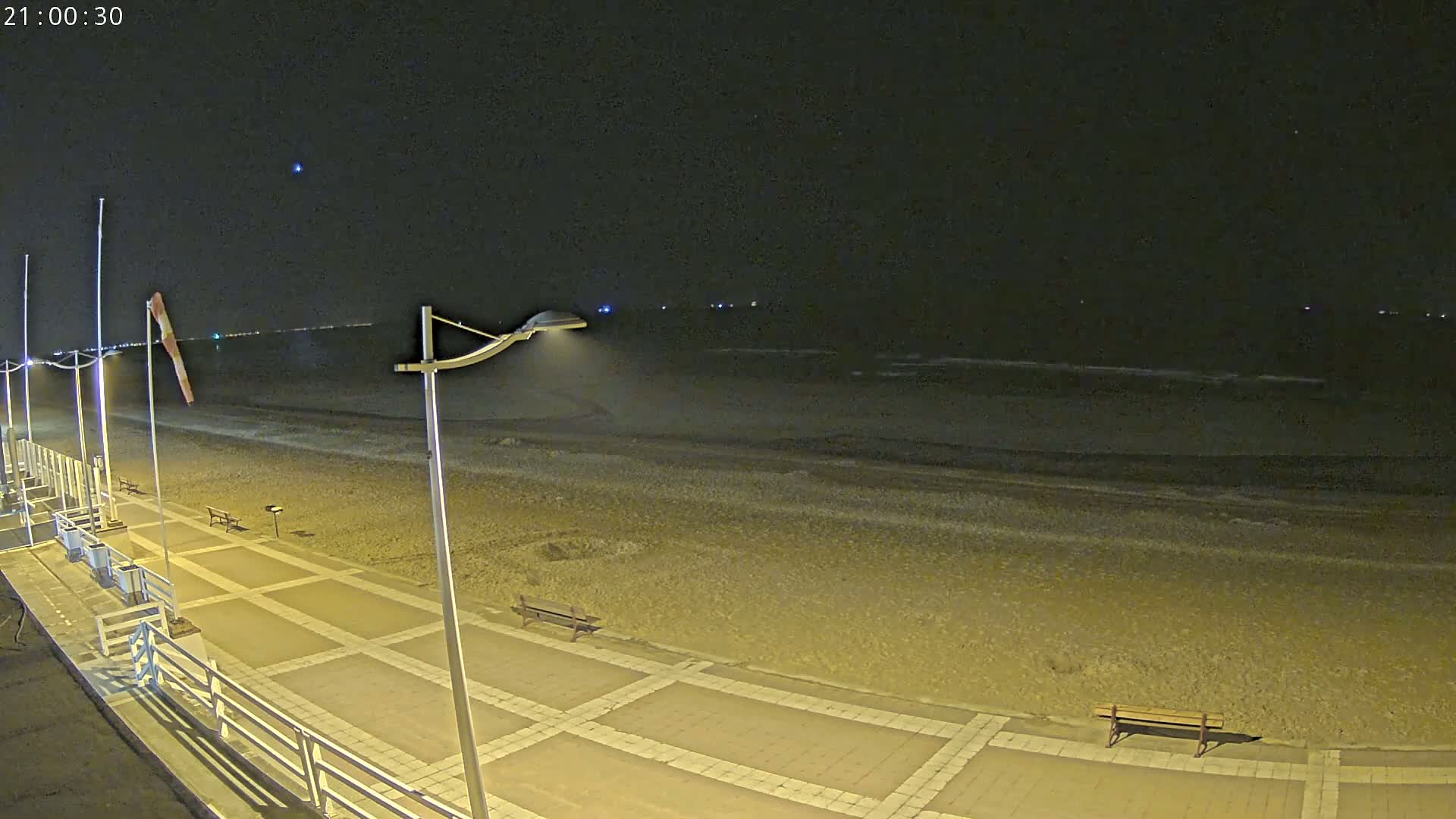 A nighttime view of a beach with a paved walkway, benches, and lights, under a clear, dark sky.