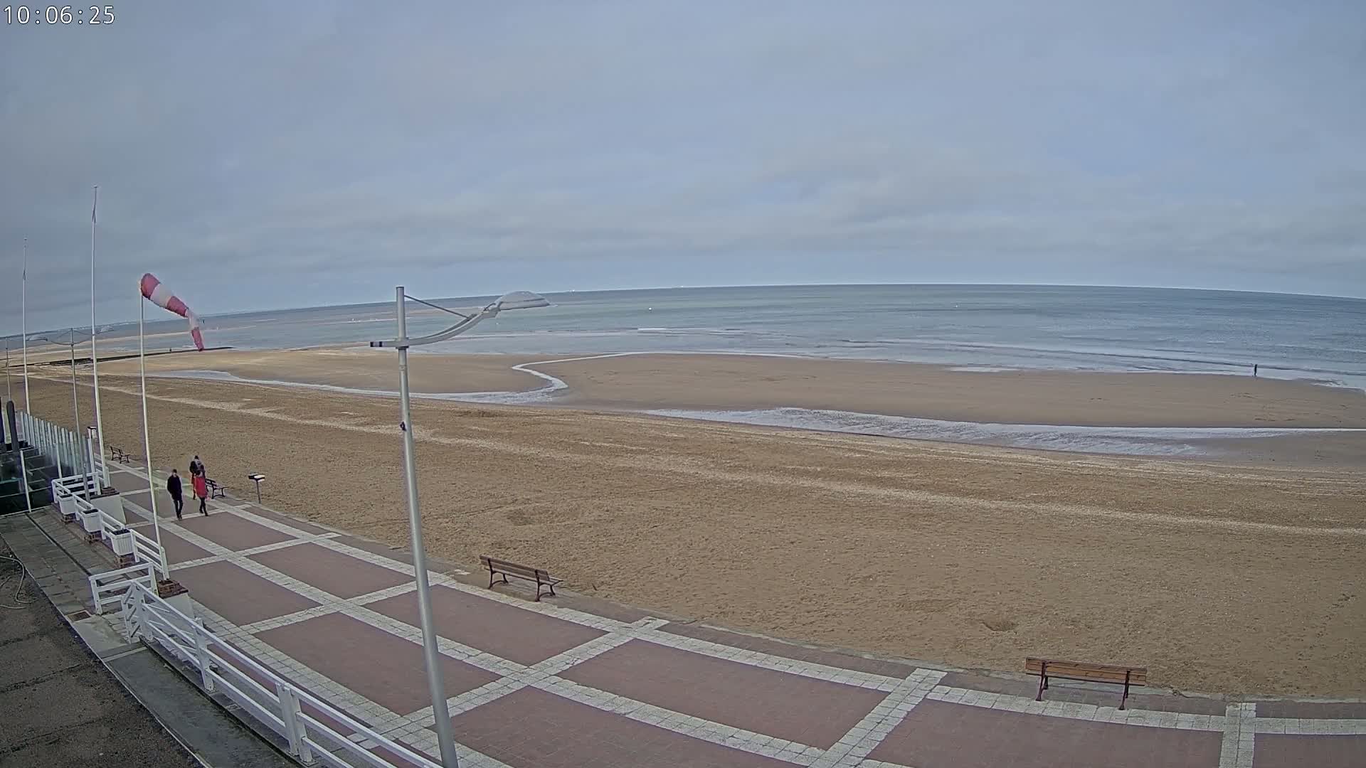 A wide shot of a mostly empty sandy beach with a paved walkway and benches, under an overcast sky with a windsock indicating wind.