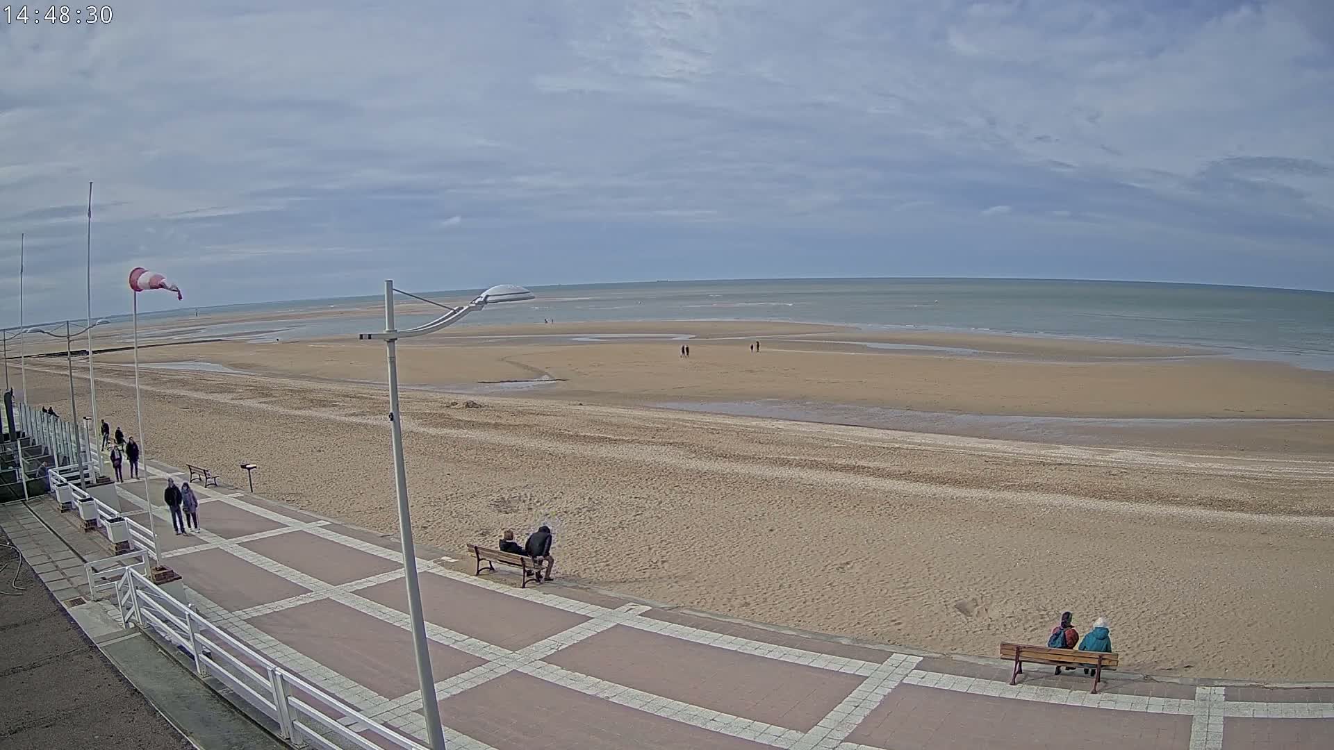 A wide sandy beach with a paved walkway, sparsely populated with people, under a partly cloudy sky.