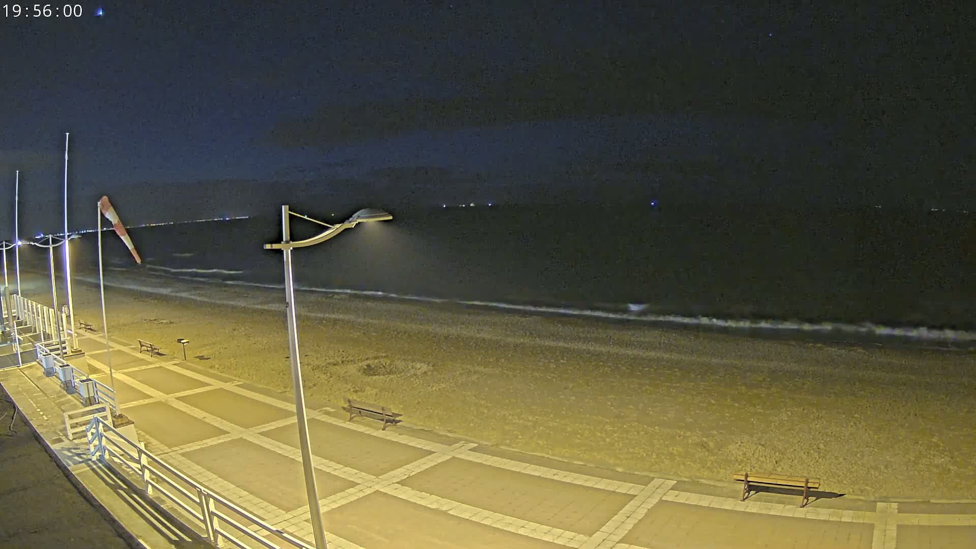 A nighttime beach scene shows a paved walkway with benches, lights, and a windsock, overlooking a dark, gently waving ocean under a clear, starry sky.