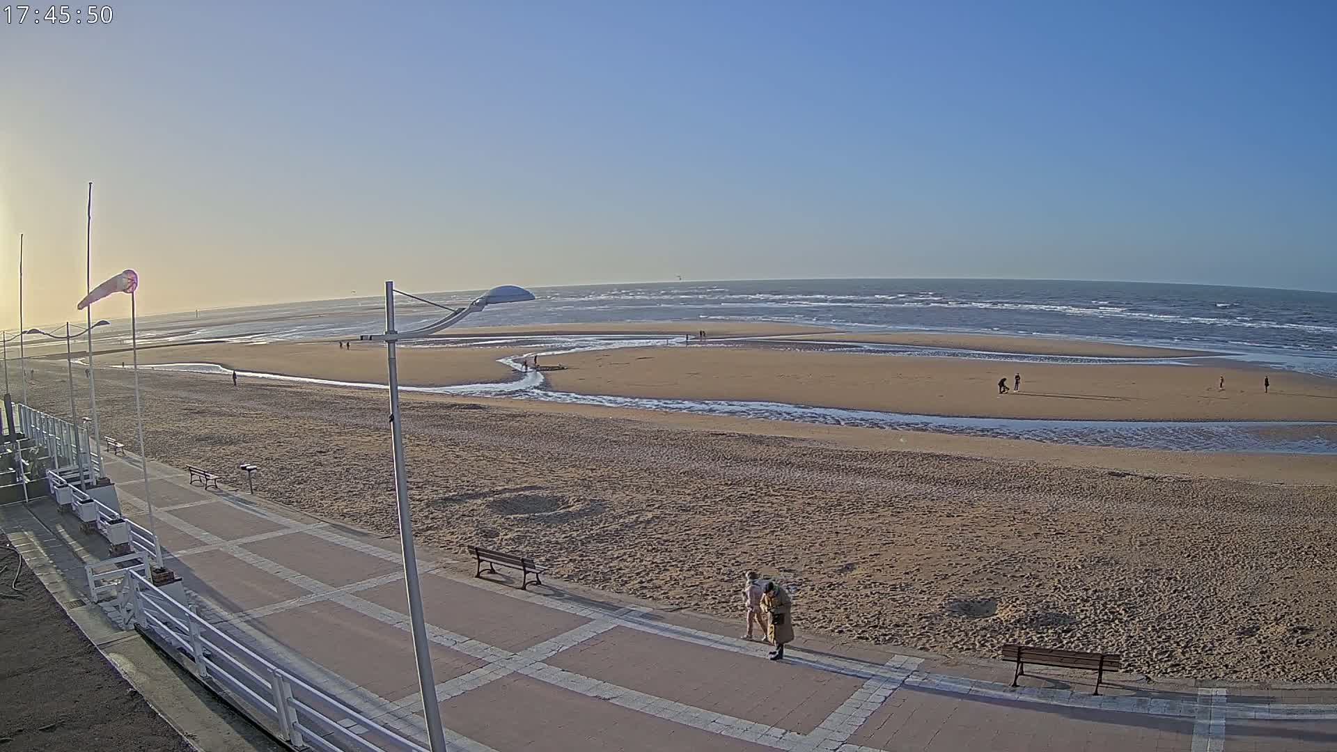 A wide sandy beach with a calm sea under a clear blue sky, showing a paved walkway with benches and a few people walking near the water's edge.