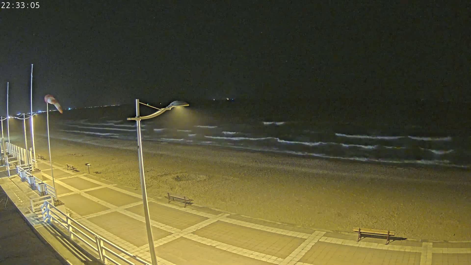 A nighttime view of a beach with gently lapping waves under a clear, dark sky, showing a paved walkway with benches and lights.
